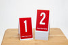 Tent style table numbers feature a bold white number on a red background. Please place visibly is printed at the bottom of each number.